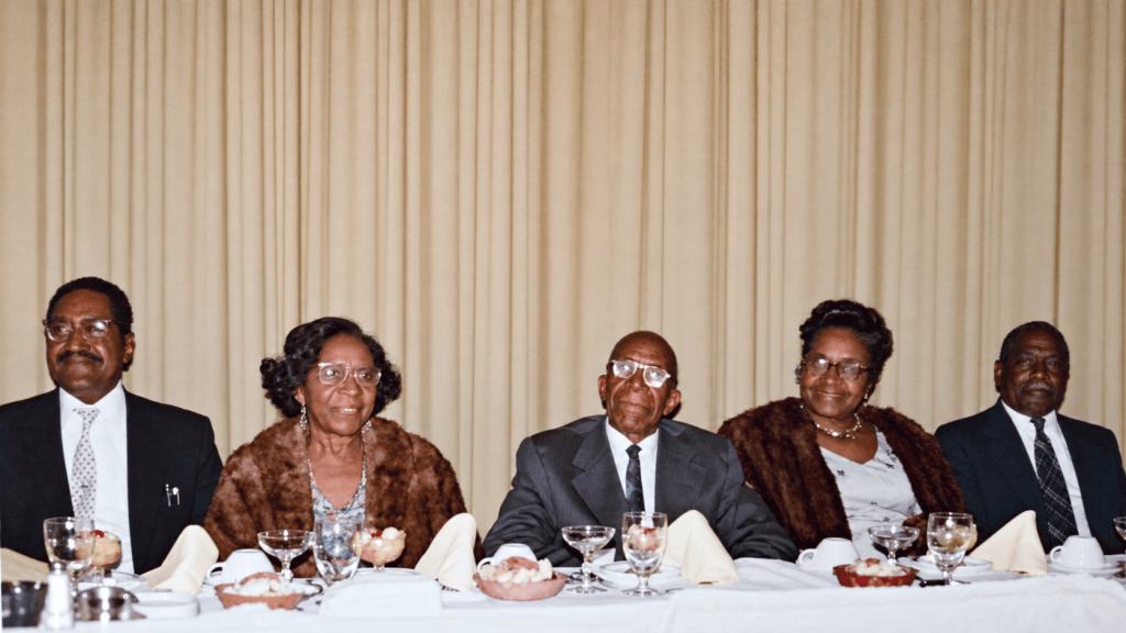 A group of five elderly individuals sitting at a dining table, dressed in formal attire, with desserts and drinks in front of them, set against a beige curtain backdrop.